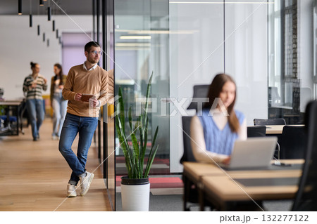 Businessman standing by glass wall in a modern office corridor, holding a digital tablet and smiling with reflection visible on the glass, symbolizing success, optimism, and modern business lifestyle. 132277122