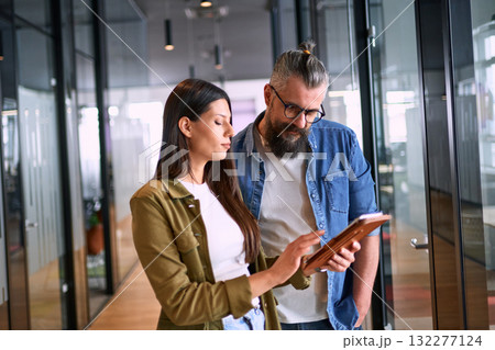 Two business professionals having a focused discussion while reviewing information on a tablet in a bright modern office hallway, symbolizing teamwork, communication, and digital collaboration. Two business professionals having a focused discussion while reviewing information on a tablet in a bright modern office hallway, symbolizing teamwork, communication, and digital collaboration. 132277124