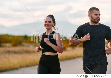 A couple dressed in sportswear runs along a scenic road during an early morning workout, enjoying the fresh air and maintaining a healthy lifestyle A couple dressed in sportswear runs along a scenic road during an early morning workout, enjoying the fresh air and maintaining a healthy lifestyle 132277246