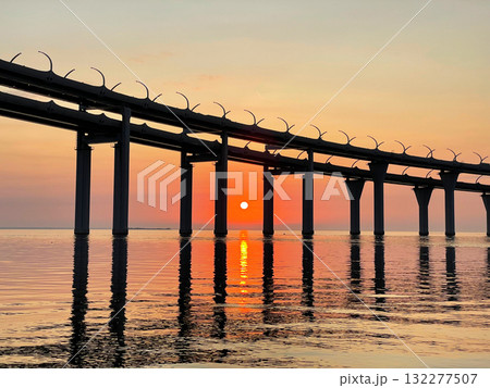 Beautiful sunset over the sea. The sun setting between the pillars of the bridge. Reflection of sunlight on water. The sky is painted in golden colors. Kanonersky Island, Saint-Petersburg Beautiful sunset over the sea. The sun setting between the pillars of the bridge. Reflection of sunlight on water. The sky is painted in golden colors. Kanonersky Island, Saint-Petersburg 132277507