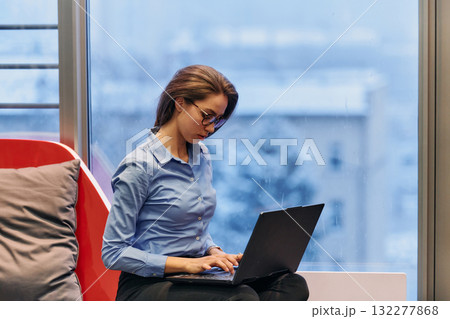 A businesswoman utilizes her laptop while seated by the window of a large corporate building, offering a picturesque view of the city skyline as her backdrop. 132277868
