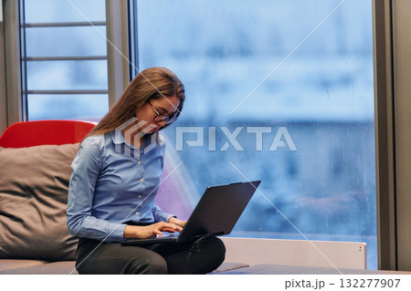 A businesswoman utilizes her laptop while seated by the window of a large corporate building, offering a picturesque view of the city skyline as her backdrop. 132277907