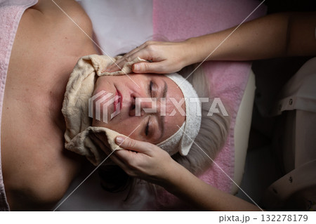 Close up of a mature woman receiving a relaxing facial massage from a professional female therapist in a modern spa. The client lies on a treatment bed with a towel and headband, enjoying skincare Close up of a mature woman receiving a relaxing facial massage from a professional female therapist in a modern spa. The client lies on a treatment bed with a towel and headband, enjoying skincare 132278179