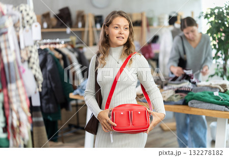 Winter collection sale. Positive young girl client standing against background of shelves with clothes in modern store and choosing red bag Winter collection sale. Positive young girl client standing against background of shelves with clothes in modern store and choosing red bag 132278182