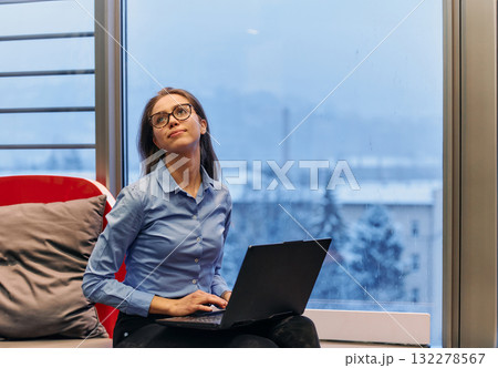 A businesswoman utilizes her laptop while seated by the window of a large corporate building, offering a picturesque view of the city skyline as her backdrop. A businesswoman utilizes her laptop while seated by the window of a large corporate building, offering a picturesque view of the city skyline as her backdrop. 132278567
