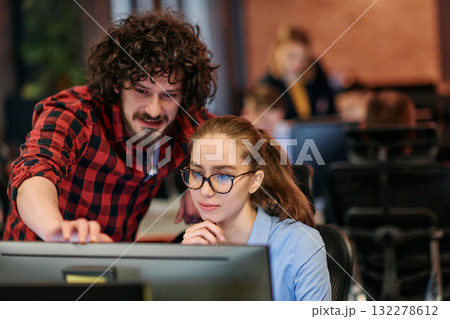 Business colleagues, a man and a woman, engage in discussing business strategies while attentively gazing at a computer monitor, epitomizing collaboration and innovation 132278612