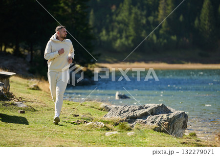 Athletic man maintains his healthy lifestyle by running through the scenic mountain and lakeside environment, showcasing a commitment to fitness and well-being Athletic man maintains his healthy lifestyle by running through the scenic mountain and lakeside environment, showcasing a commitment to fitness and well-being 132279071