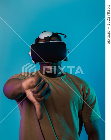 African American man, wearing virtual reality glasses, expresses dissatisfaction by pointing down with his hand while standing isolated against a blue background, portraying a modern critique within 132279251