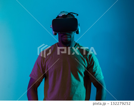 In a futuristic visual, an African American man stands isolated against a striking blue backdrop, adorned with VR glasses that transport him into a cutting-edge virtual reality experience, merging 132279252
