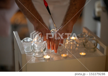 Close up view of a therapists hands preparing glass cups and lighting candles for traditional fire cupping therapy. Concept of alternative medicine, relaxation, and natural healing treatments. 132279286
