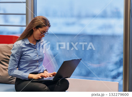 A businesswoman utilizes her laptop while seated by the window of a large corporate building, offering a picturesque view of the city skyline as her backdrop. 132279600