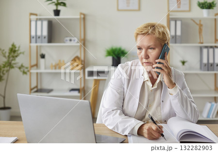 Doctor at laptop, senior medical mature woman using smartphone, input information in copy book Doctor at laptop, senior medical mature woman using smartphone, input information in copy book 132282903