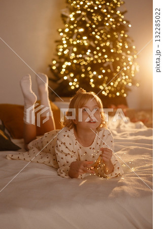 Smiling girl in white dress lying on bed with Christmas tree lights in cozy room Smiling girl in white dress lying on bed with Christmas tree lights in cozy room 132285022