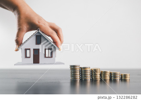 Hand holding house model above stacked coins, symbolizing mortgage loan, real estate investment, property finance, savings, wealth growth, home ownership and housing economy on minimal background. 132286282