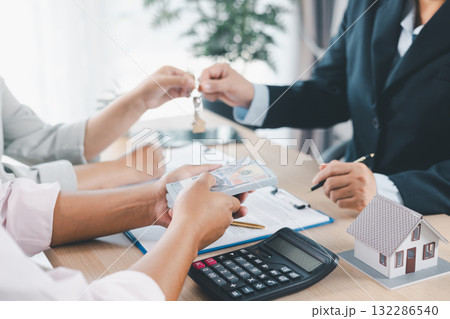 Close up of real estate agent handing house key to client with contract paperwork, calculator and house models on desk, concept of home ownership, mortgage loan, property investment and success. Close up of real estate agent handing house key to client with contract paperwork, calculator and house models on desk, concept of home ownership, mortgage loan, property investment and success. 132286540