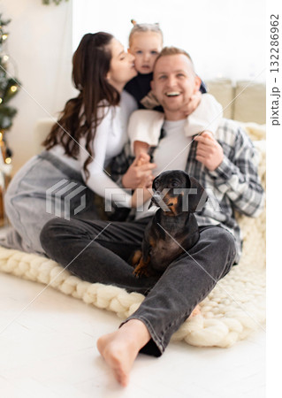 A happy family of three and their dachshund dog are sitting together indoors. The mother is kissing the child on the cheek. A happy family of three and their dachshund dog are sitting together indoors. The mother is kissing the child on the cheek. 132286962