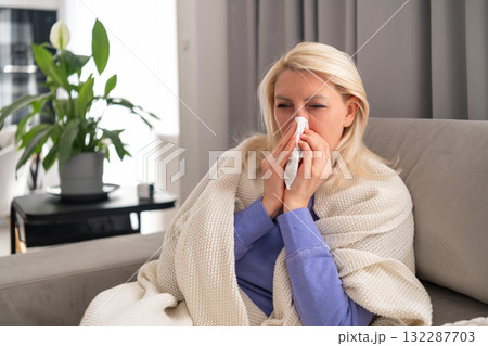 Ill woman covered with a warm plaid sitting on sofa and blowing her nose with tissue. Concept of flu, cold, home treatment, and seasonal illness during epidemic. 132287703