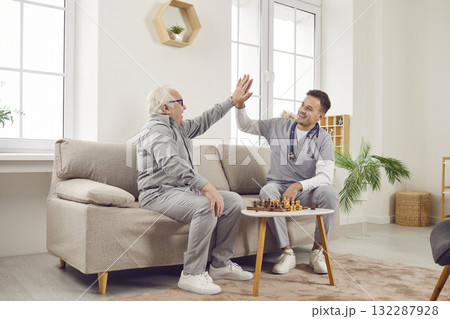 Nurse man giving high five to a gray-haired senior elderly person while playing chess game at home. Nurse man giving high five to a gray-haired senior elderly person while playing chess game at home. 132287928