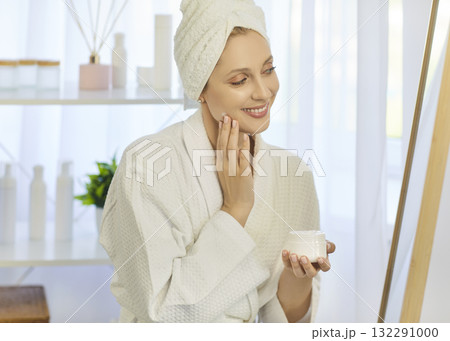Blonde woman applying face cream at home in towel and robe during skincare routine Blonde woman applying face cream at home in towel and robe during skincare routine 132291000