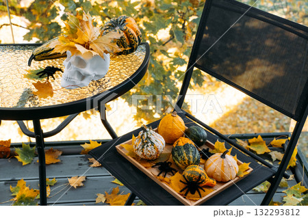 White decorative skull with maple leaves and mixed gourds on a glass table and chair on autumn balcony. Modern spooky decor, visual Halloween culture, alternative seasonal styling, aesthetic horror 132293812