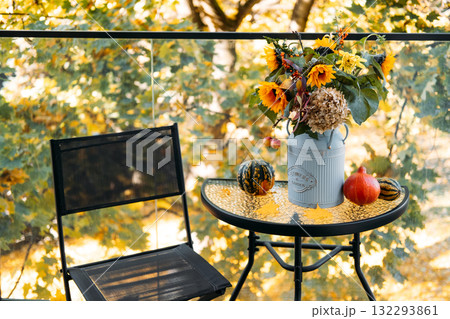 Simple autumn balcony setup with floral centerpiece in a vintage can beside gourds and a mesh chair against fall foliage. Compact seasonal design, minimalist outdoor decor, cozy urban living Simple autumn balcony setup with floral centerpiece in a vintage can beside gourds and a mesh chair against fall foliage. Compact seasonal design, minimalist outdoor decor, cozy urban living 132293861