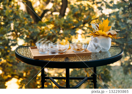 Outdoor glass table setup featuring a skull adorned with autumn leaves next to a wooden tray filled with assorted supplements. Mortality and health awareness in autumn visuals, memento mori, 132293865