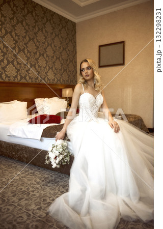 Blonde bride sitting on bed in white dress with bouquet in hands Blonde bride sitting on bed in white dress with bouquet in hands 132298231