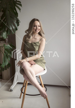An elegant blonde girl in a thin dress sits on a chair in a minimalist interior. An elegant blonde girl in a thin dress sits on a chair in a minimalist interior. 132298258