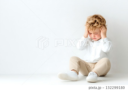 A young boy sits on a white floor holding his head in a pensive worried expression conveying a sense of distress vulnerability and inner struggle A young boy sits on a white floor holding his head in a pensive worried expression conveying a sense of distress vulnerability and inner struggle 132299180