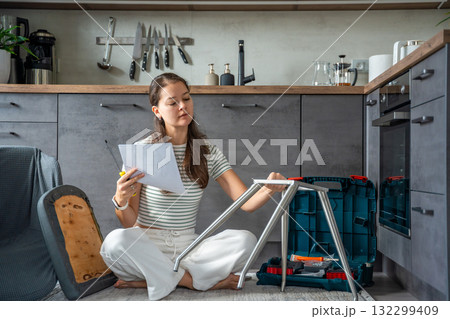 Smiling young woman sitting on the floor reading instructions while assembling a chair. Continuation of the series showing joy and satisfaction from independent DIY work at home. Smiling young woman sitting on the floor reading instructions while assembling a chair. Continuation of the series showing joy and satisfaction from independent DIY work at home. 132299409