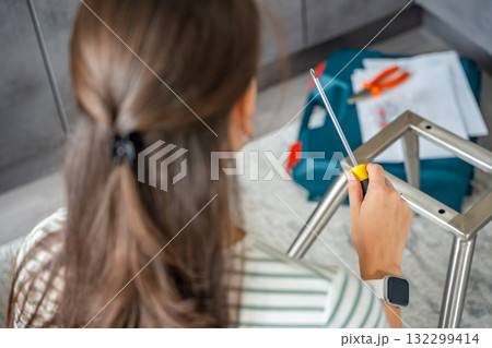 Young woman seen from behind assembling a chair with screwdrivers on the floor at home. Concept of modern lifestyle and practical skills in everyday life. Young woman seen from behind assembling a chair with screwdrivers on the floor at home. Concept of modern lifestyle and practical skills in everyday life. 132299414
