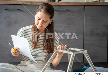 Smiling young woman sitting on the floor reading instructions while assembling a chair. Continuation of the series showing joy and satisfaction from independent DIY work at home. Smiling young woman sitting on the floor reading instructions while assembling a chair. Continuation of the series showing joy and satisfaction from independent DIY work at home. 132299429