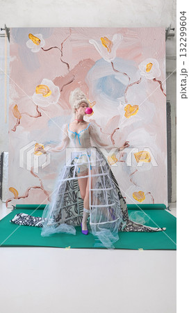 Playful woman blowing pink bubblegum in front of floral backdrop in character of Marie Antoinette. Playful woman blowing pink bubblegum in front of floral backdrop in character of Marie Antoinette. 132299604