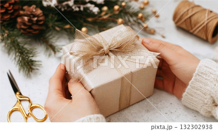 Female hands carefully wrap a small gift box in cozy decorations, preparing for a joyful Christmas celebration. Female hands carefully wrap a small gift box in cozy decorations, preparing for a joyful Christmas celebration. 132302938
