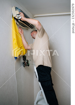 Vertical shot of skilled HVAC professional standing on ladder and disassembling wall-mounted air conditioning unit, using yellow drainage bag while carefully accessing electrical components Vertical shot of skilled HVAC professional standing on ladder and disassembling wall-mounted air conditioning unit, using yellow drainage bag while carefully accessing electrical components 132302979