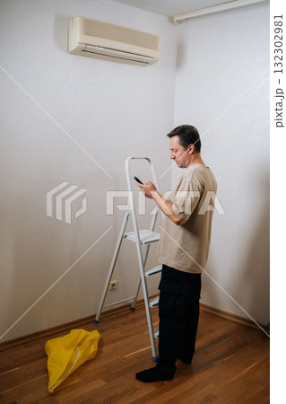 Vertical shot of technician male standing by ladder, using smartphone app to control and prepare for air conditioner maintenance, ensuring optimal cooling performance and indoor air quality. Vertical shot of technician male standing by ladder, using smartphone app to control and prepare for air conditioner maintenance, ensuring optimal cooling performance and indoor air quality. 132302981