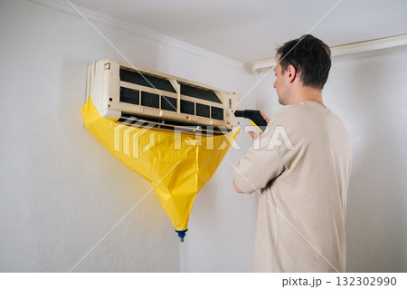 Air conditioning technician cleaning air conditioner unit using steam cleaner. While yellow protective bag collects dirty water. Ensuring thorough and professional cleaning process. 132302990