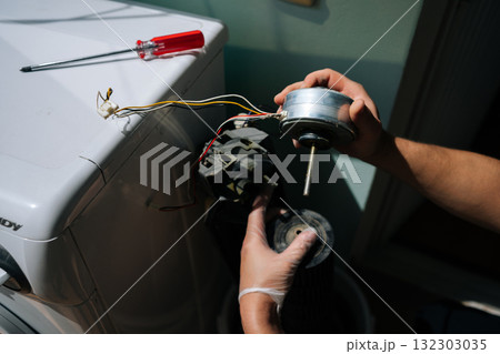 Close-up hands of unrecognizable technician wearing safety gloves examines components of disassembled washing machine, focusing on detached motor while troubleshooting malfunction. 132303035