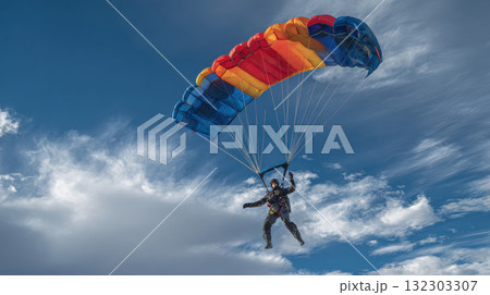 Skydiver with colorful parachute soaring through dramatic cloudy sky, extreme sport adventure with vibrant red, orange and blue canopy against clouds 132303307