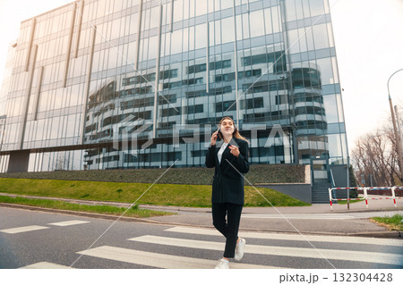 Young Woman Walks Confidently Across a Pedestrian Crosswalk in an Urban Setting During Daylight, Reflecting Modern Architecture of Nearby Buildings 132304428