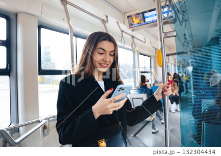 Young Woman Enjoys Texting on Her Smartphone While Riding a City Tram in the Afternoon, Surrounded by Fellow Commuters and Urban Scenery 132304434