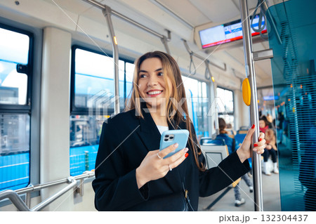 Smiling Young Woman Inside Modern Urban Tram, Holding Smartphone While Enjoying the City Ambiance on a Sunny Day Smiling Young Woman Inside Modern Urban Tram, Holding Smartphone While Enjoying the City Ambiance on a Sunny Day 132304437
