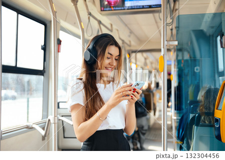 Young Woman Smiles While Using Smartphone on Public Transport With Headphones During Daytime Commute Young Woman Smiles While Using Smartphone on Public Transport With Headphones During Daytime Commute 132304456