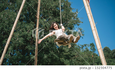 Joyful child swinging on a wooden swing in a sunny outdoor park surrounded by green trees with blue sky above, capturing happiness and freedom 132304775