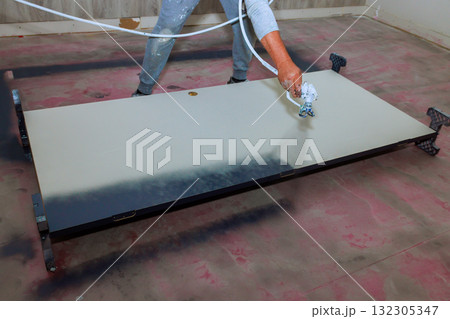 Worker applies spray gun paint to white door while renovating room 132305347