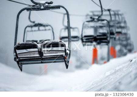 Snow-covered ski lift chairs hanging in the air above a wintery landscape during a snowy day at the mountain resort 132306430