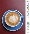 beautifully crafted latte with intricate foam art sits in blue cup on white saucer, against rich maroon background, evoking sense of warmth and comfort 132308825