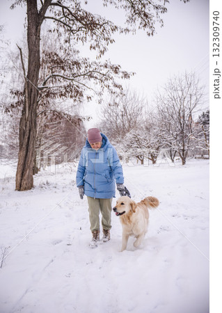 Elderly woman in a blue winter coat walking her Golden Retriever through a snowy rural landscape. The dog walks happily beside her as she enjoys a quiet moment outdoors surrounded by winter trees. Elderly woman in a blue winter coat walking her Golden Retriever through a snowy rural landscape. The dog walks happily beside her as she enjoys a quiet moment outdoors surrounded by winter trees. 132309740