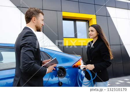 Sales manager showing how to charge electric car to a young client woman, plugging wire into the car socket at the car dealership outdoor Sales manager showing how to charge electric car to a young client woman, plugging wire into the car socket at the car dealership outdoor 132310170