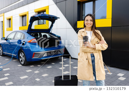 Young woman with cup of coffee waiting while her electric car charging, sustainable and economic transportation concept Young woman with cup of coffee waiting while her electric car charging, sustainable and economic transportation concept 132310179